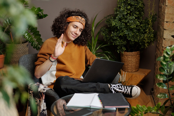 A young man with black curly hair in a bandana is sitting on a chair with his legs crossed with a laptop on his lap and smiling communicating via video link in a room with a lot of plants